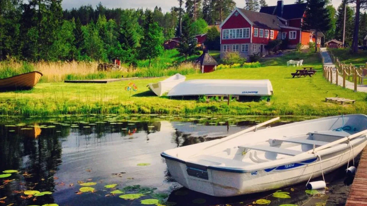 Odalgårdens gräsmatta och röda byggnader vid sjön, med roddbåtar uppdragna vid vattenbrynet en sommardag.