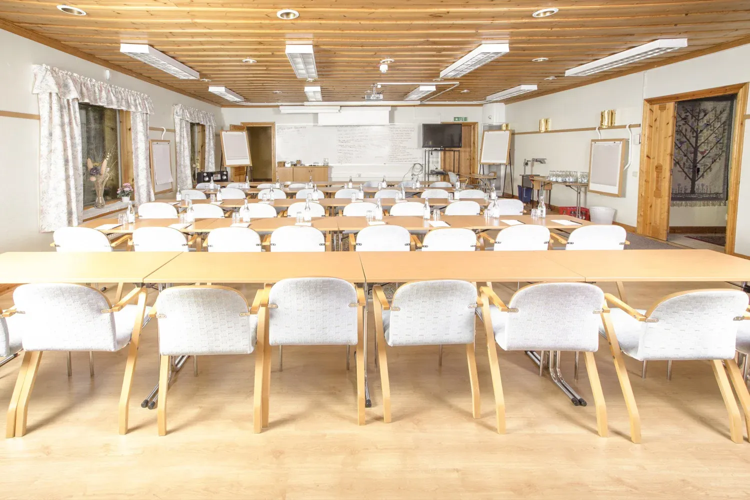 Konferenslokal uppställd med långa bord, vita stolar och dagsljus från fönsterraden.