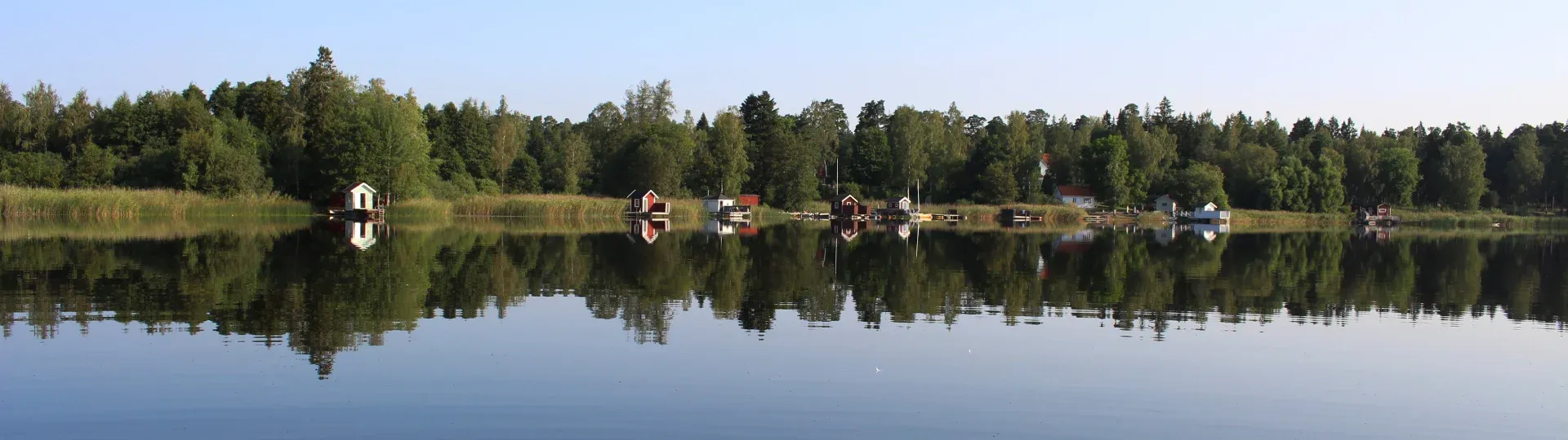 Sjön spegelblank med skogsklädd strand och byggnader längs vattenlinjen.