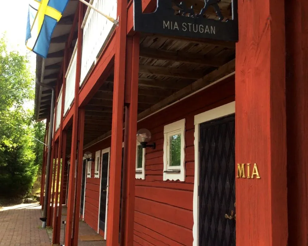 Röd stugbyggnad med vita fönsterfoder och två svenska flaggor vid entrén.